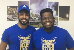 Antônio Gonzaga e André Rodrigues posam de pé contra uma parede com fotos de desfiles da Portela atrás