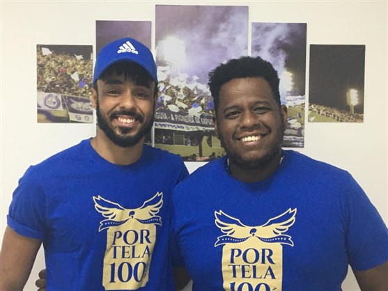 Antônio Gonzaga e André Rodrigues posam de pé contra uma parede com fotos de desfiles da Portela atrás